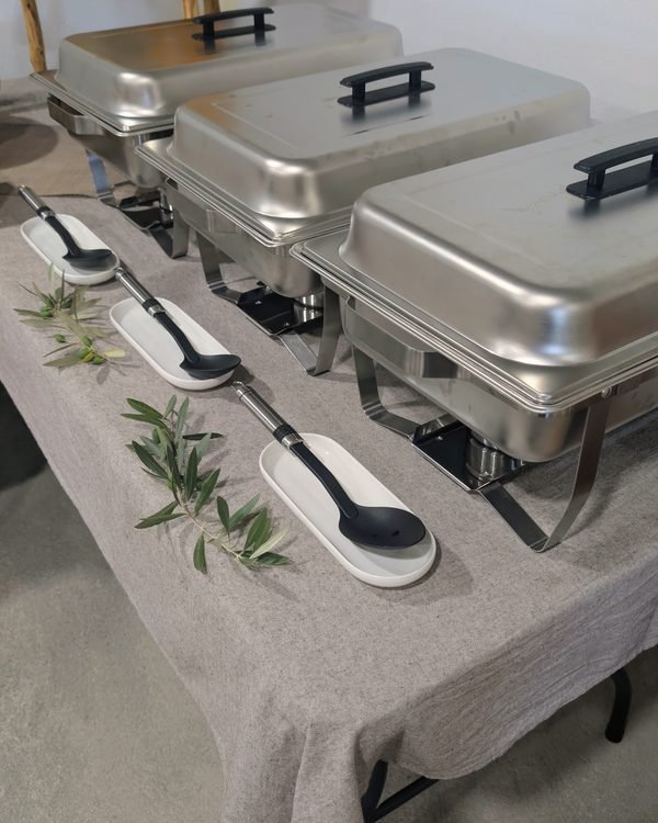 Mesa de buffet com réchauds e ramos de oliveira sobre toalha de linho